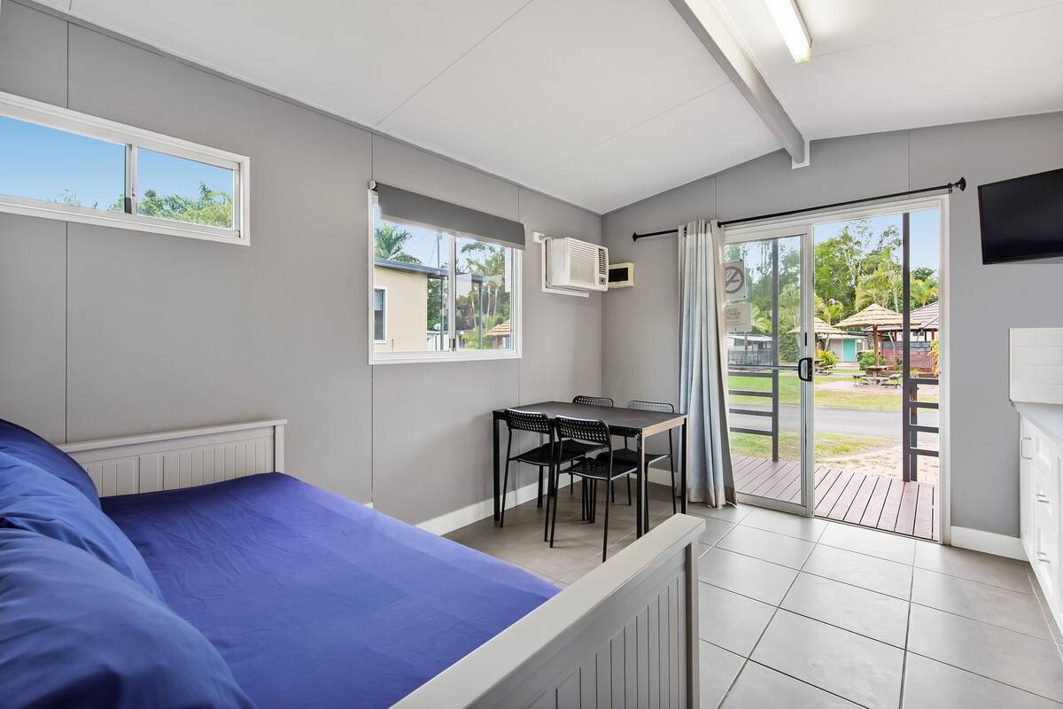 Una habitación con una cama, una mesa con sillas, un ventanal y una puerta que da a un espacio exterior.
