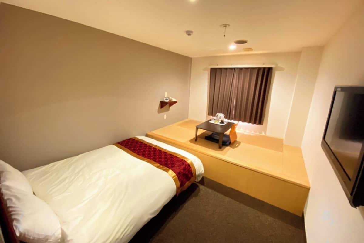 A hotel room with a single bed, a small table with a chair, a TV mounted on the wall, and a window with curtains.