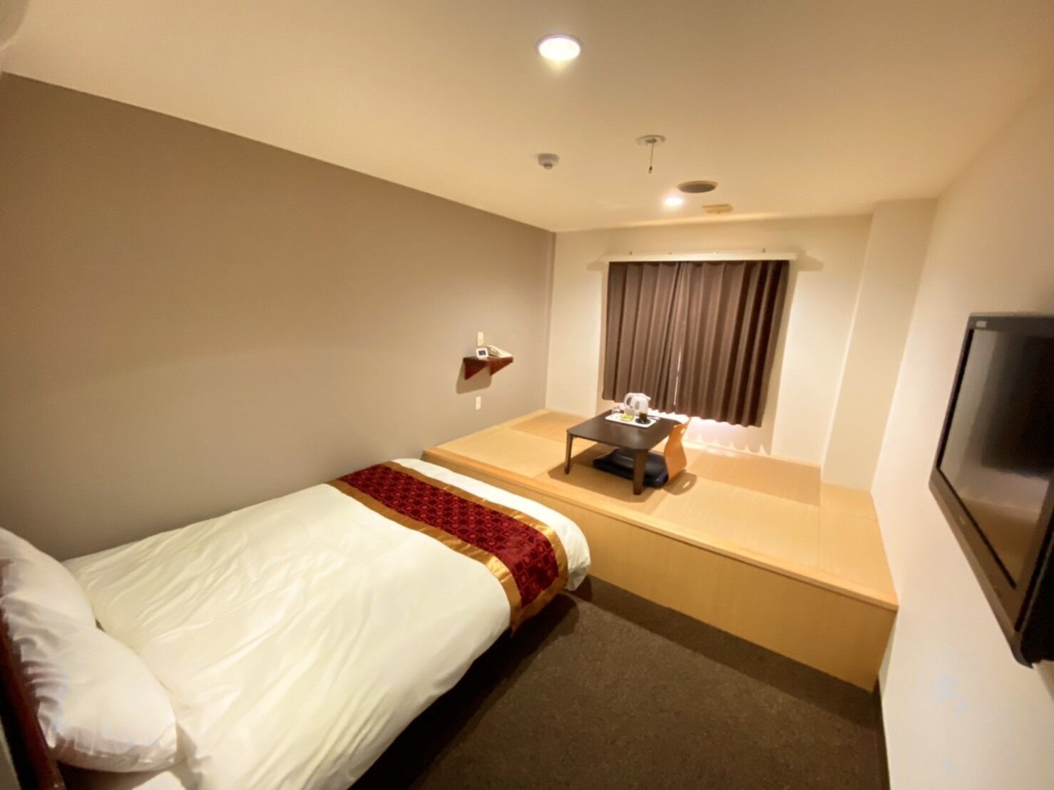 Ein Hotelzimmer mit einem Einzelbett, einem kleinen Tisch mit Wasserkocher, einem Fernseher und einem Fenster mit Vorhängen.