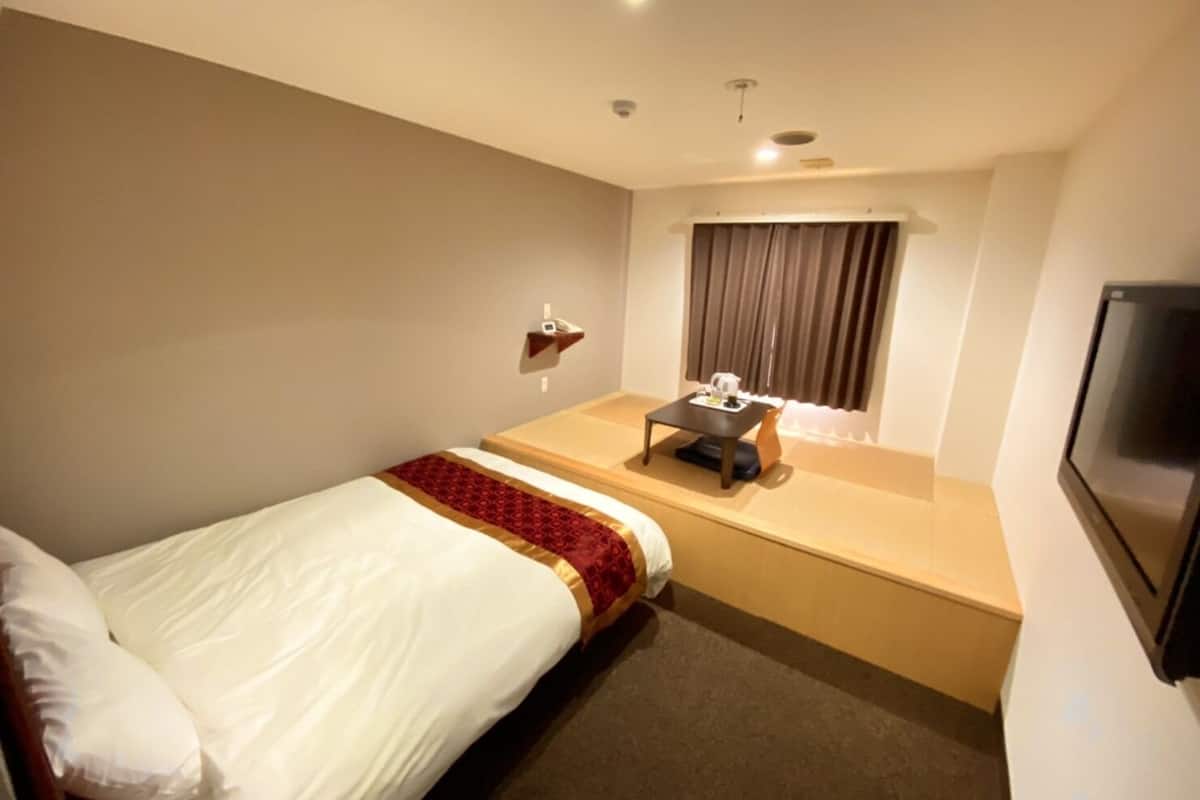 A hotel room with a single bed, a small table with a kettle, a TV, and a window with curtains.