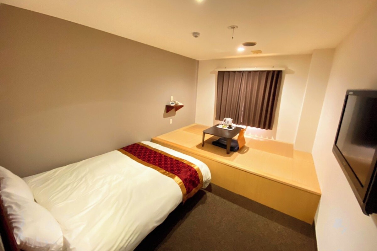 A hotel room with a single bed, a small table with a chair, a TV mounted on the wall, and a window with curtains.