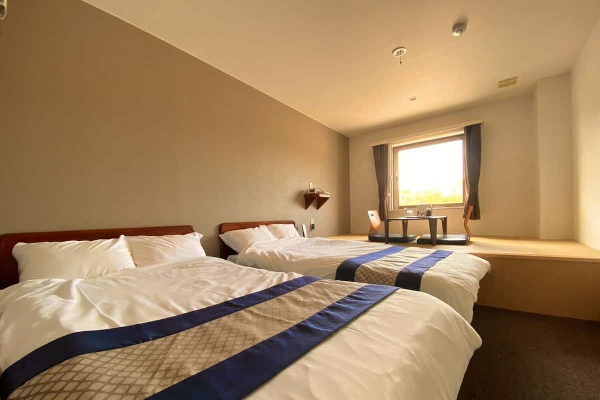 A hotel room with two beds, a window with curtains, a small table, and a wall-mounted lamp.