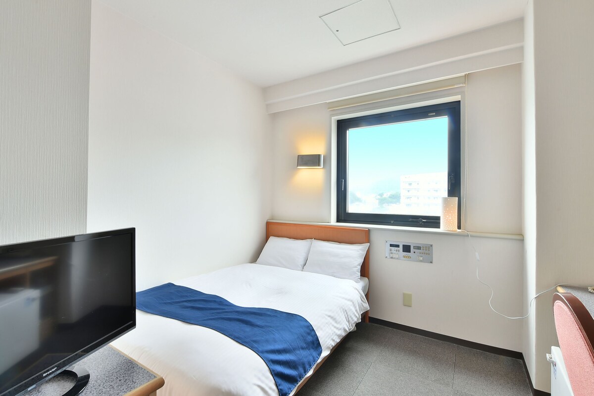 A hotel room with a bed, a TV, a window, and a control panel on the wall.