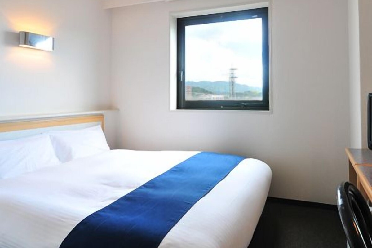A hotel room with a bed, a window with a view, and a wall-mounted light fixture.