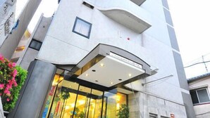 A modern building with a glass entrance, steps leading up to it, and a small garden area with pink flowers. - First Inn Haiki (Sasebo)