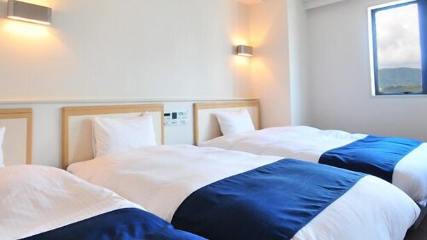 A hotel room with two beds, each with a wooden headboard, white bedding, and a blue accent. There are wall-mounted lights on either side of the beds. - First Inn Haiki (Sasebo)