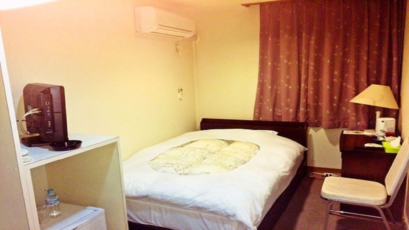 Une chambre d’hôtel comprenant un lit, un bureau avec un ordinateur, une chaise, un climatiseur et une fenêtre avec des rideaux.