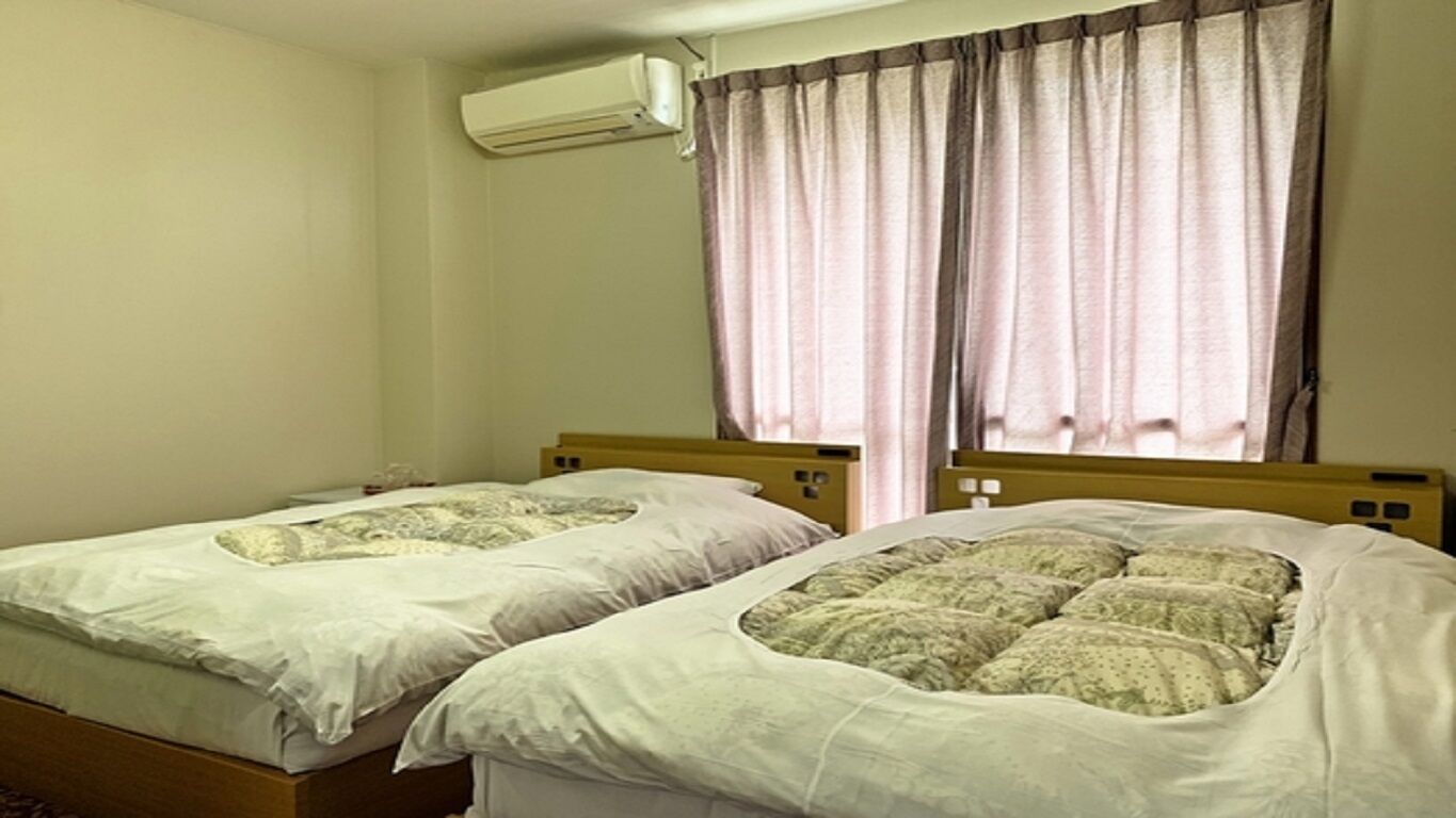 A room with two single beds, a window with curtains, an air conditioning unit, and a plain wall.