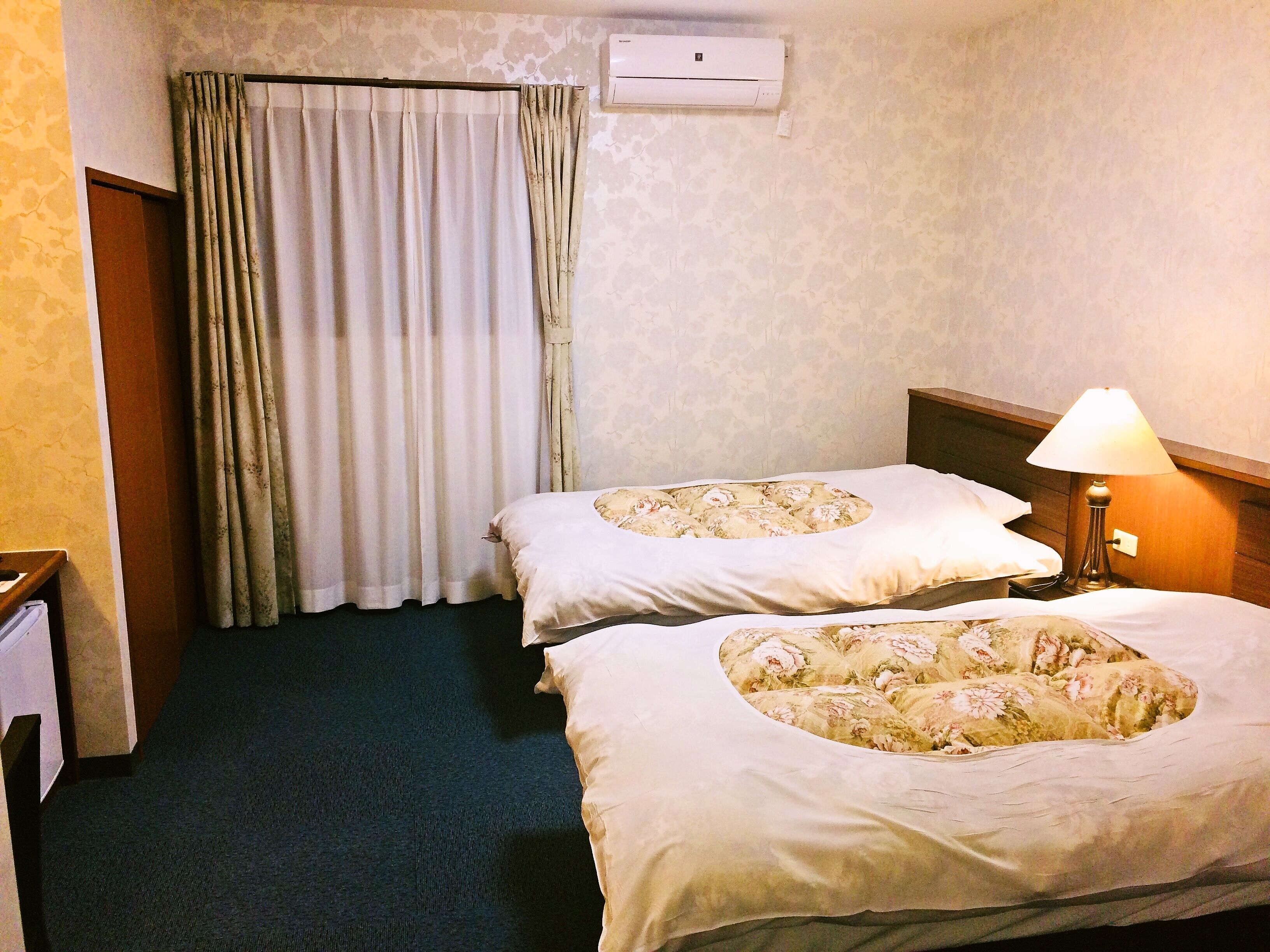 A hotel room with two beds, a curtain, an air conditioning unit, a lamp, and a wooden headboard.