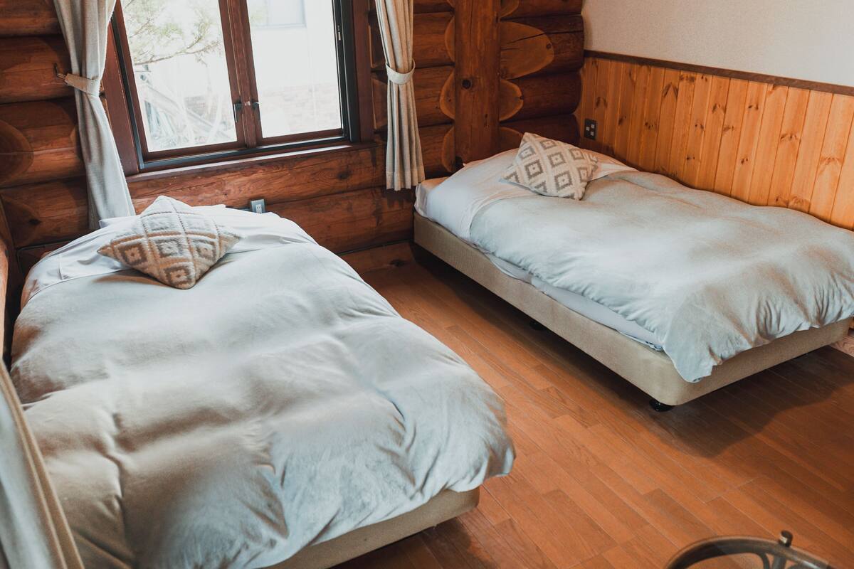 A cabin with two beds, wooden walls, a window with curtains, and a wooden floor.