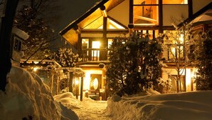 A snow-covered landscape with a large, illuminated building featuring a prominent triangular roof and multiple windows. - Hakuba Pension Meteor (Hakuba)