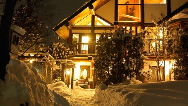 A snow-covered landscape with a large, illuminated building featuring a prominent triangular roof and multiple windows. - Hakuba Pension Meteor (Hakuba)