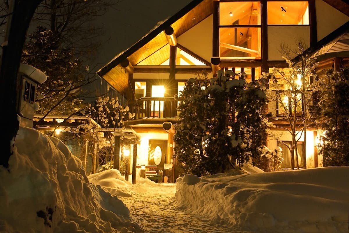 A snow-covered landscape with a large, illuminated building featuring a prominent triangular roof and multiple windows.