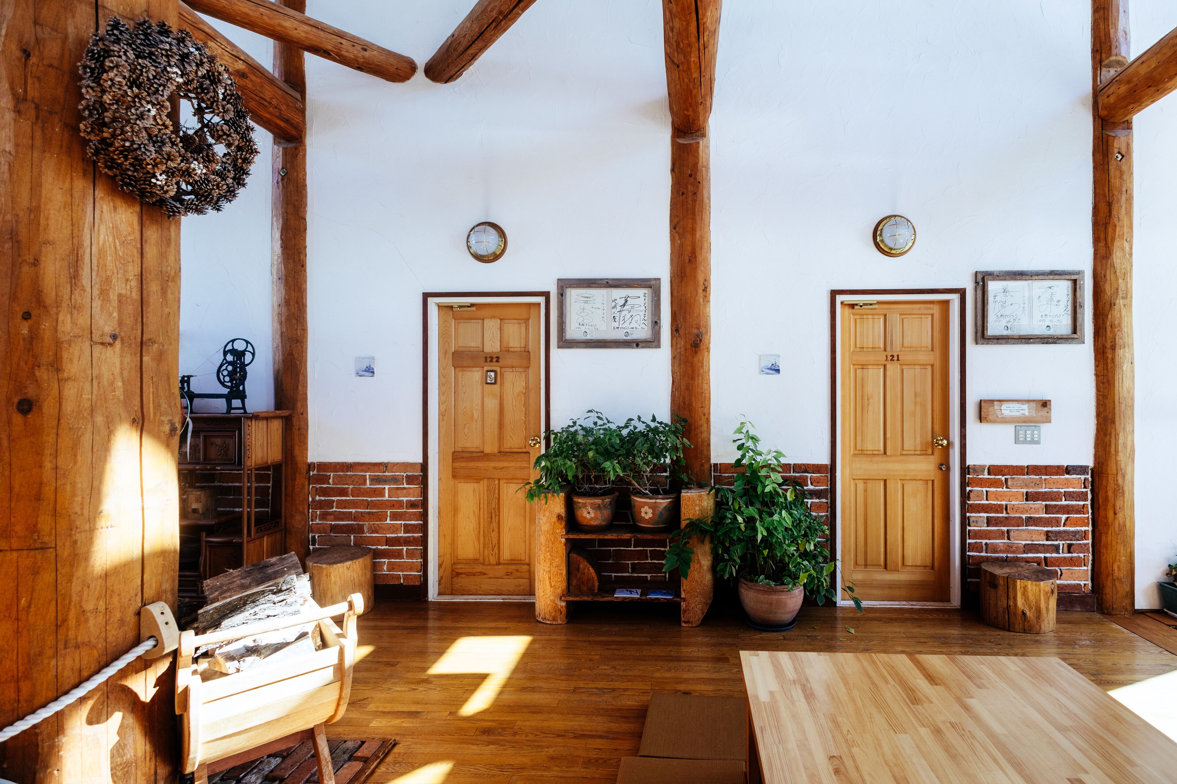 A rustic interior with wooden beams, a brick wall, and wooden doors. There are potted plants and a wooden bench.