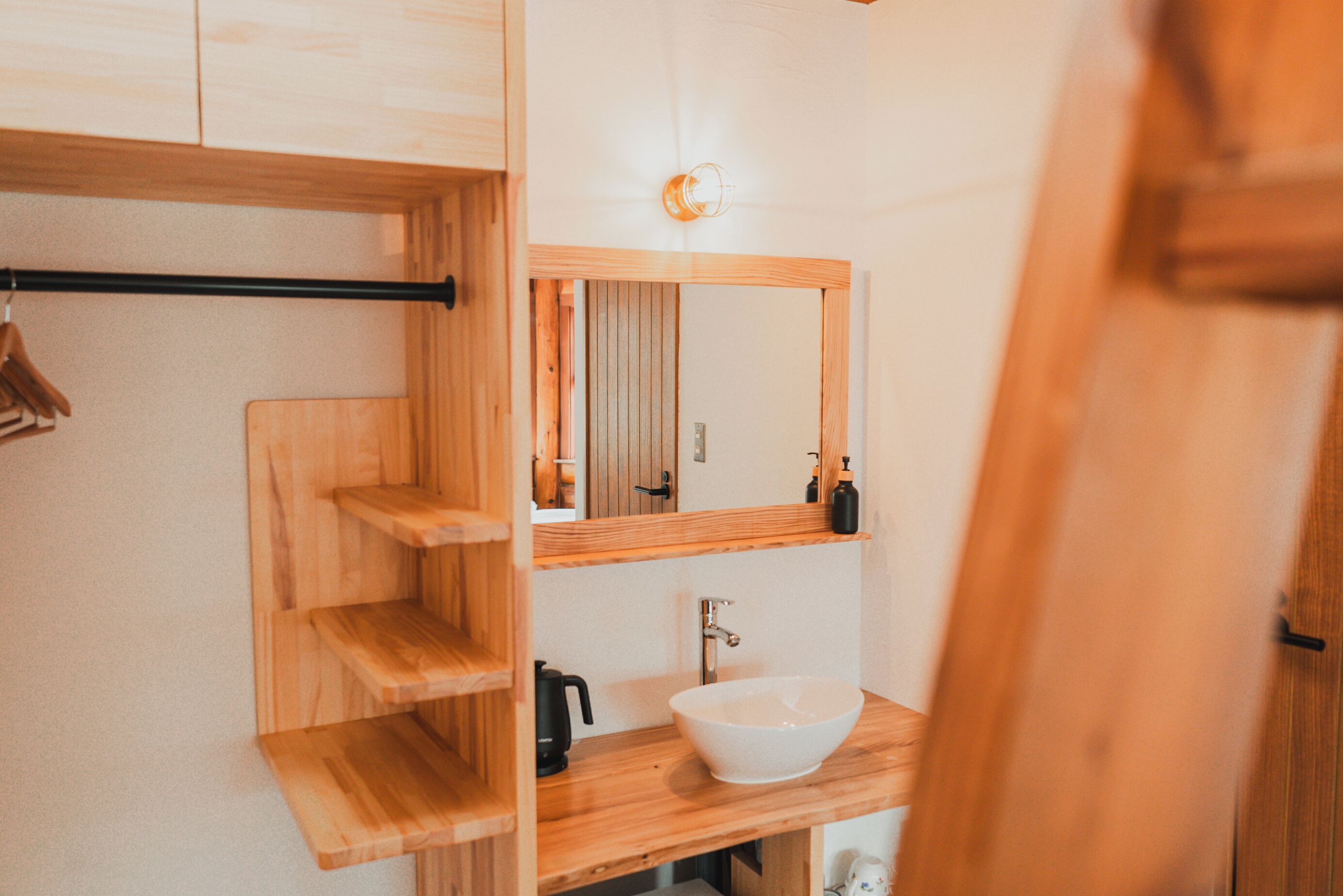 A wooden vanity with a large mirror, a sink, and a shelf with a black electric kettle.