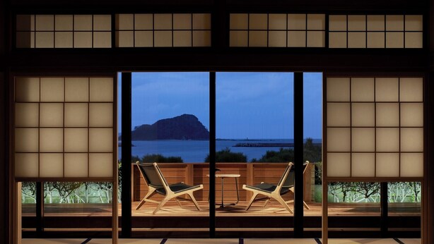 Ein traditioneller japanischer Raum mit Tatami-Boden, einem niedrigen Tisch und zwei Stühlen. Ein großes Fenster bietet Blick auf ein Gewässer und eine entfernte Insel.