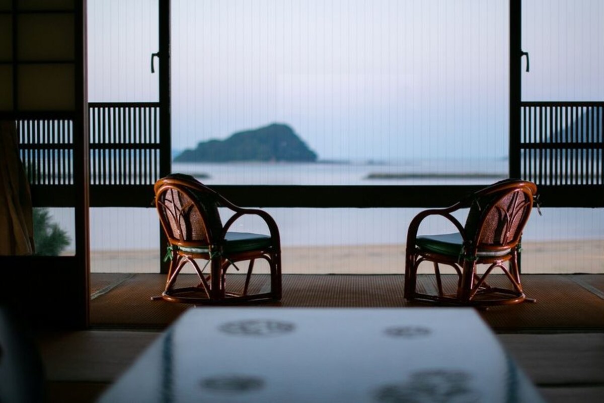 Una habitación con dos sillas de madera orientadas hacia una ventana grande que da a una playa y una isla distante.