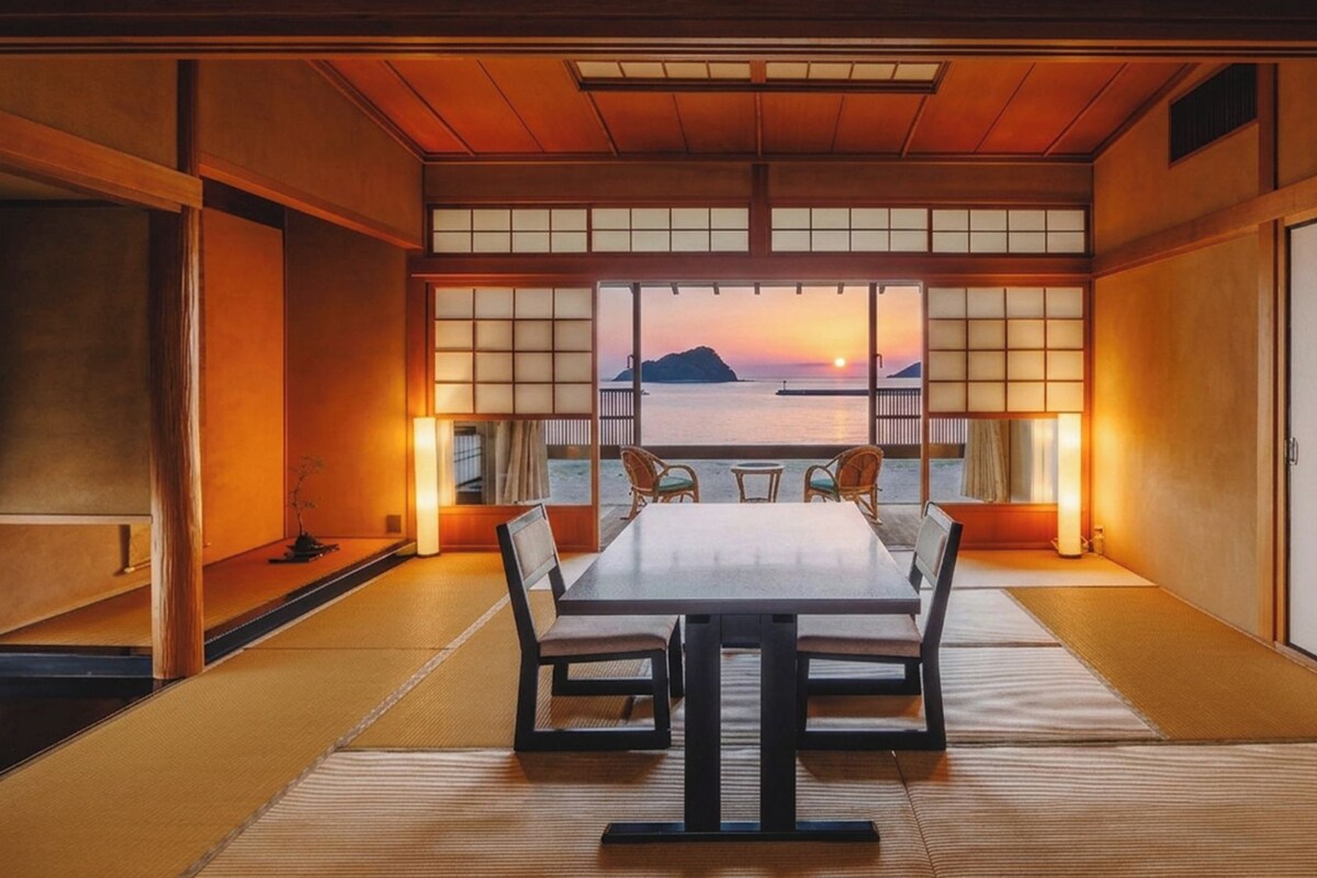 A traditional Japanese room with tatami flooring, a low table, and chairs, featuring a view of the sunset over the sea.