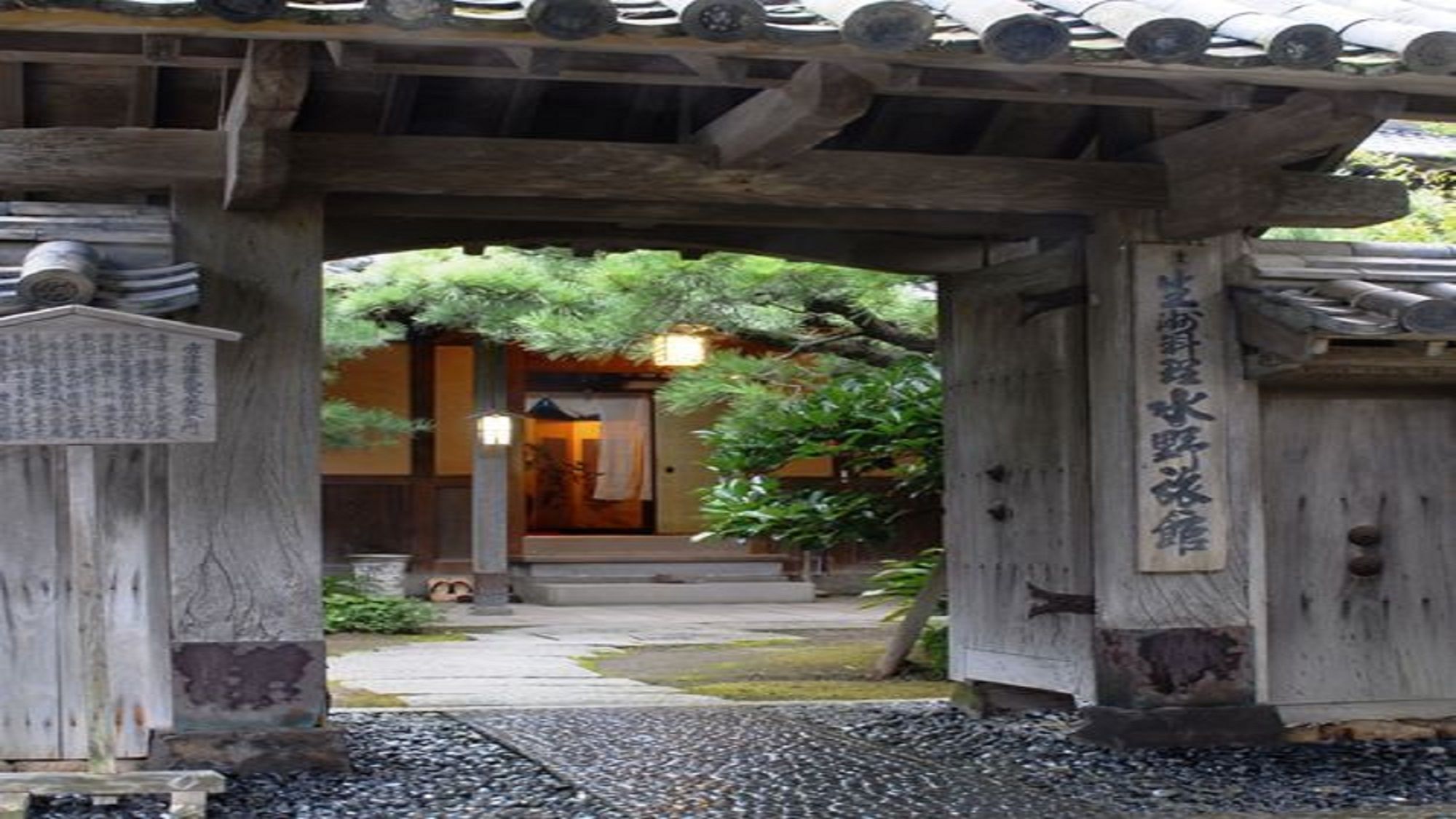 A traditional wooden structure with a covered entrance, a stone pathway, and a sign with Japanese characters.