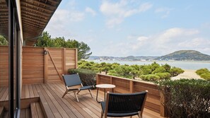 Una terraza de madera con dos sillas y una mesa pequeña, con vista a una playa y un paisaje urbano.