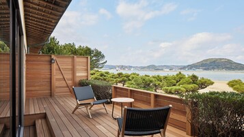 Una terraza de madera con dos sillas y una mesa pequeña, con vista a una playa y un paisaje urbano.