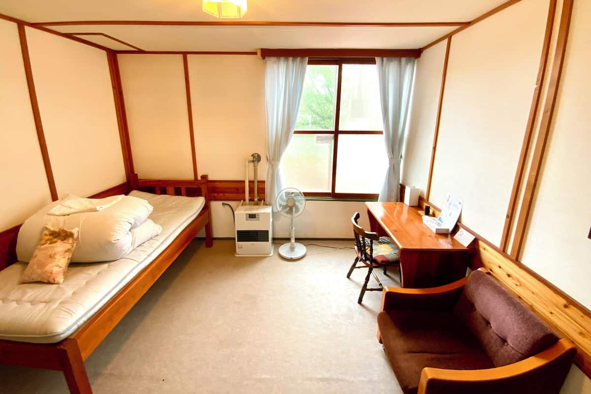A small, cozy room with a bed, a desk, a chair, and a fan.