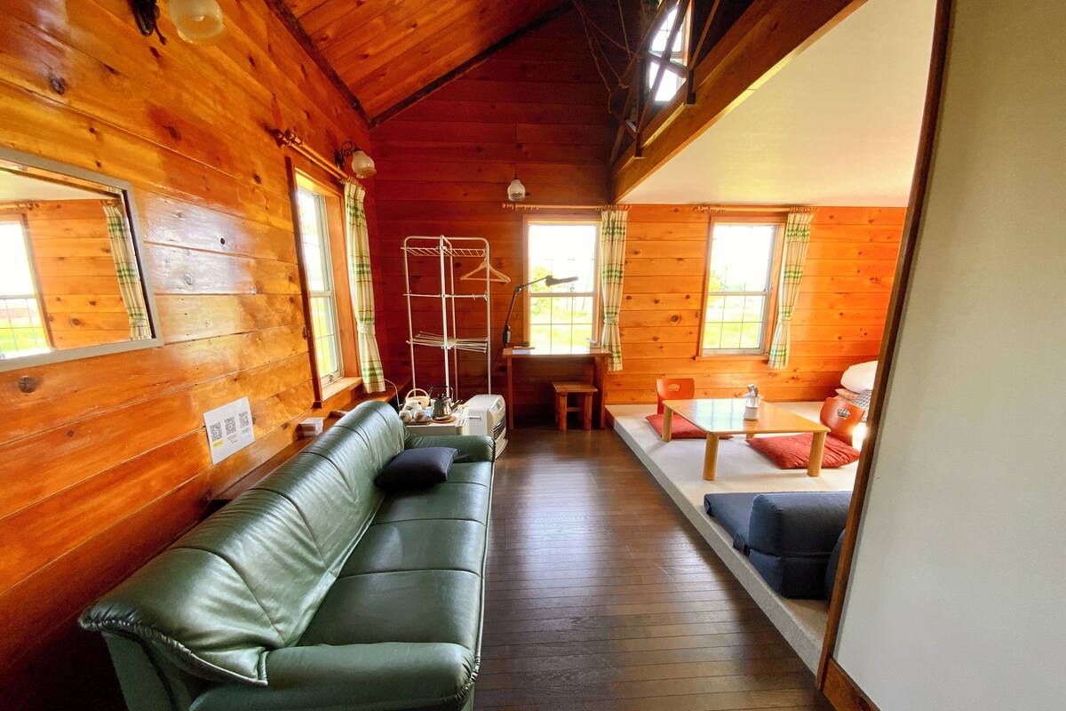 A wooden cabin interior with a green leather sofa, a dining table, and a small kitchen area.