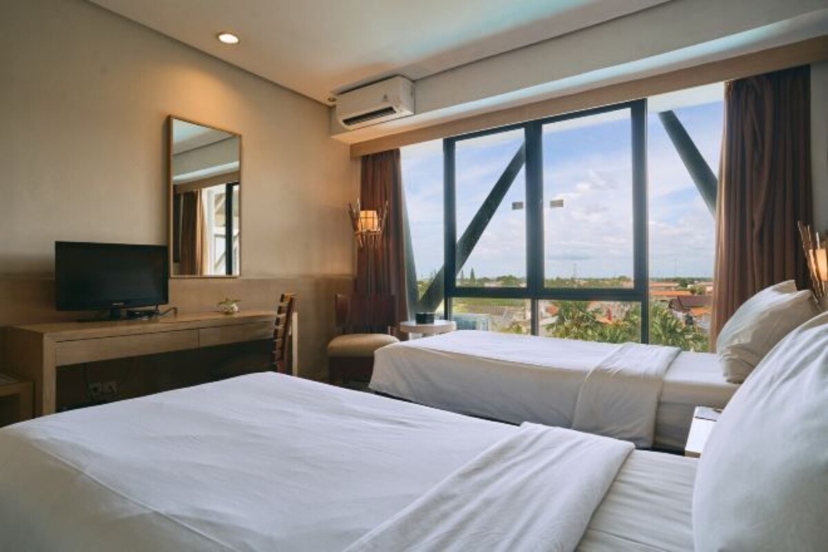 A hotel room with two beds, a TV, a desk, and a large window offering a city view.
