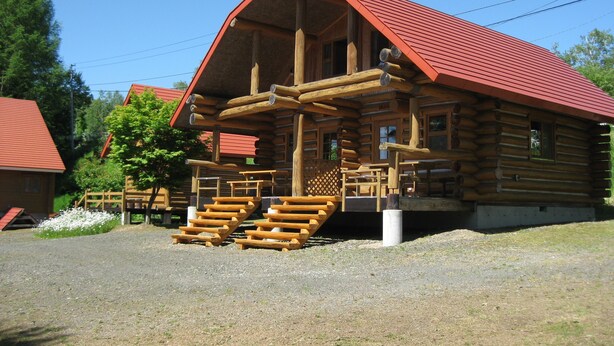A log cabin with a red roof, wooden exterior, and a porch with benches. - Log Cottage Himawari (Nakafurano)
