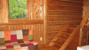 A log cabin interior with a wooden floor, a colorful patchwork sofa, a wooden ladder, and a window with a view of trees. - Log Cottage Himawari (Nakafurano)