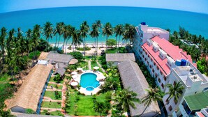 An aerial view of a resort with a swimming pool surrounded by palm trees, a beach, and hotel buildings.