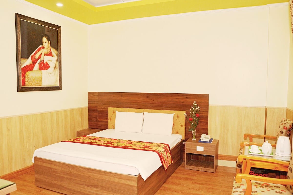 A hotel room with a wooden bed, a bedside table with a phone and a vase of flowers, a chair, and a painting on the wall.