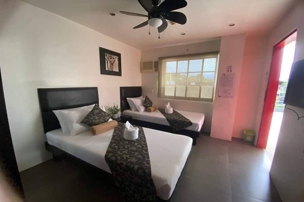 A hotel room with two beds, a ceiling fan, a window with blinds, and a red door.