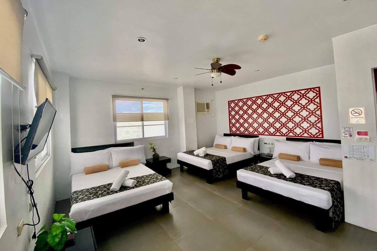 A hotel room with three beds, a ceiling fan, a TV, and a decorative red wall panel.
