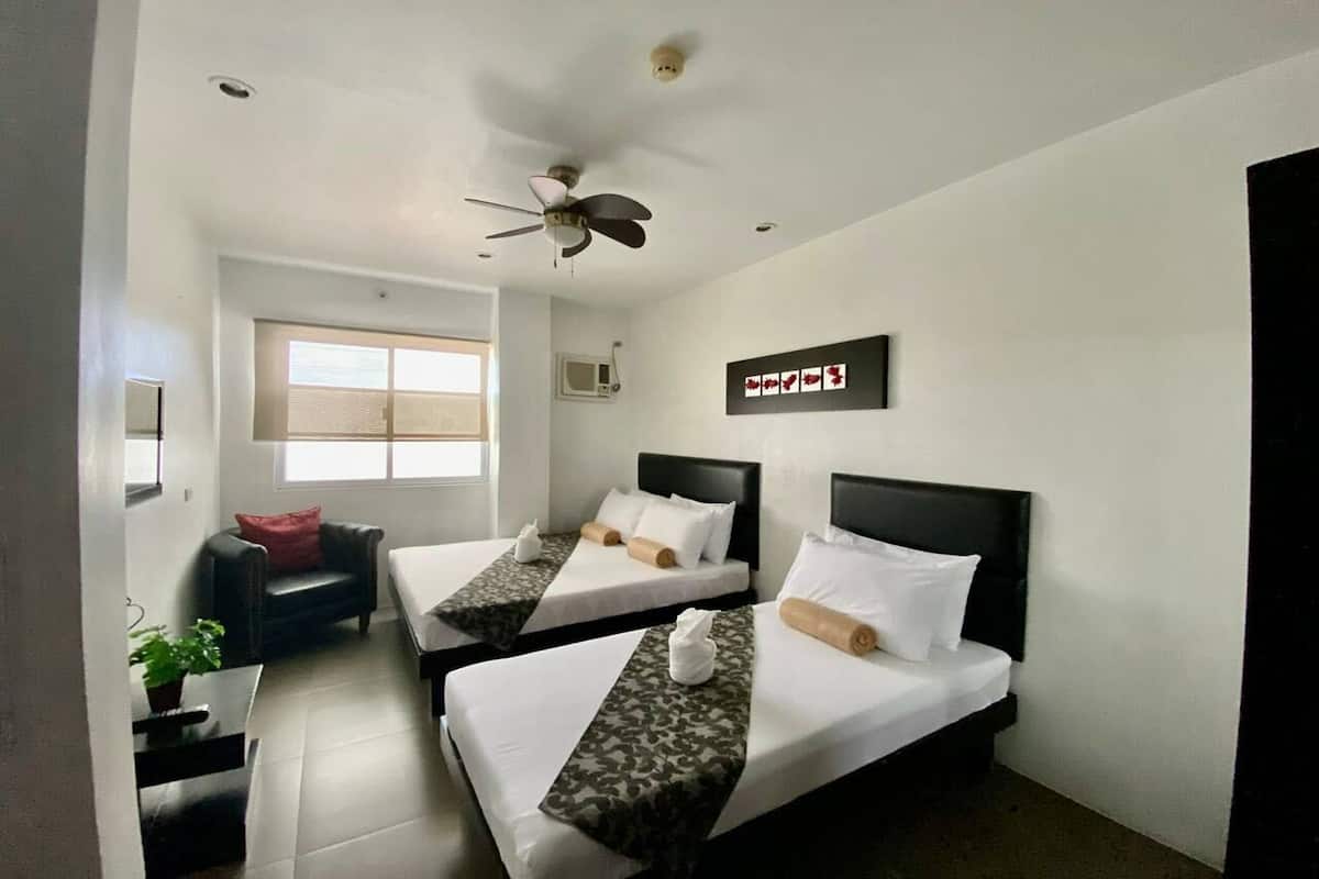 A hotel room with two beds, a ceiling fan, a window with blinds, a small table with a plant, and a wall-mounted sign.