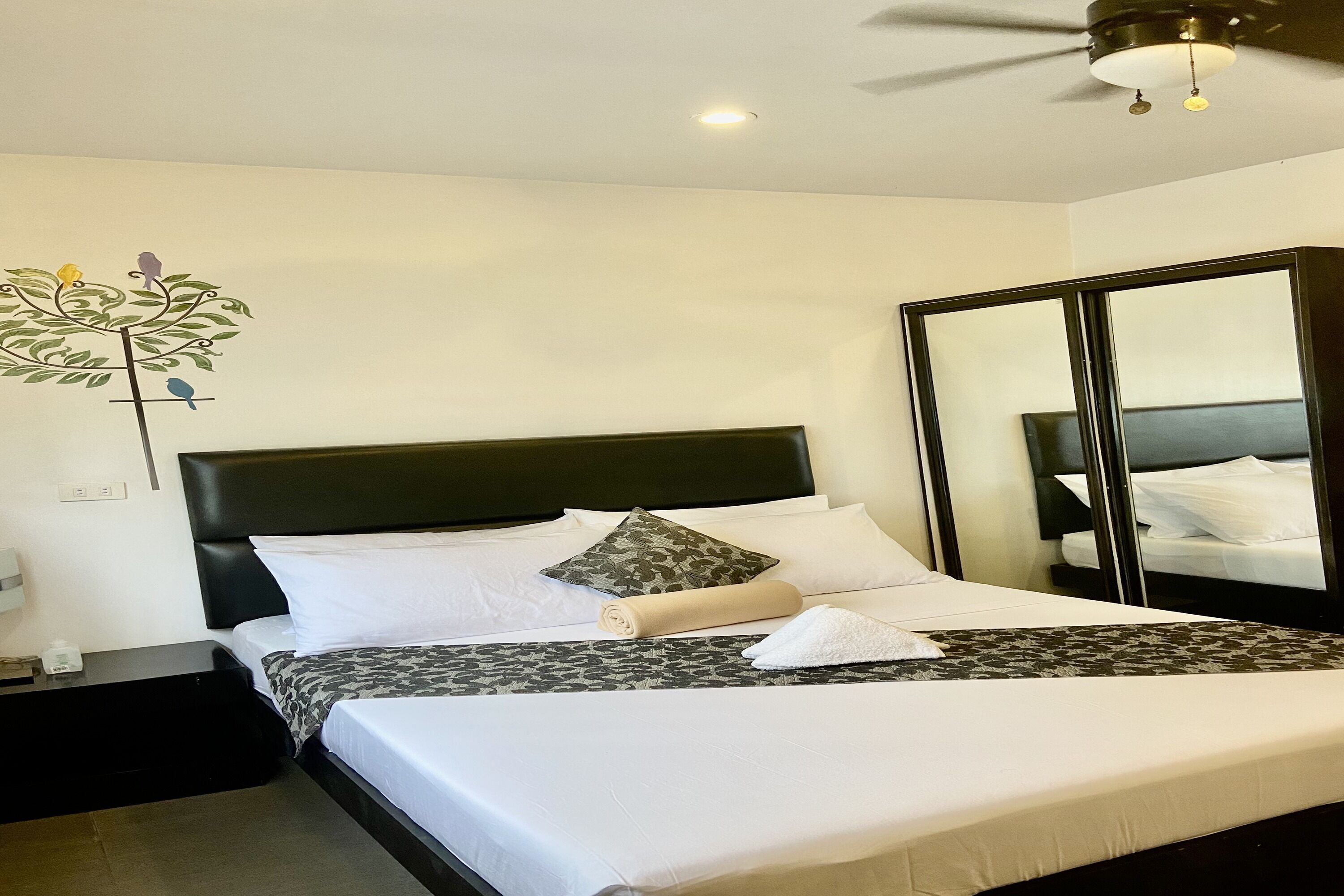 A neatly made bed with white linens, a patterned bedspread, and a rolled towel. There is a nightstand with a lamp and a small table beside the bed. A large mirror is mounted on the wall to the right.