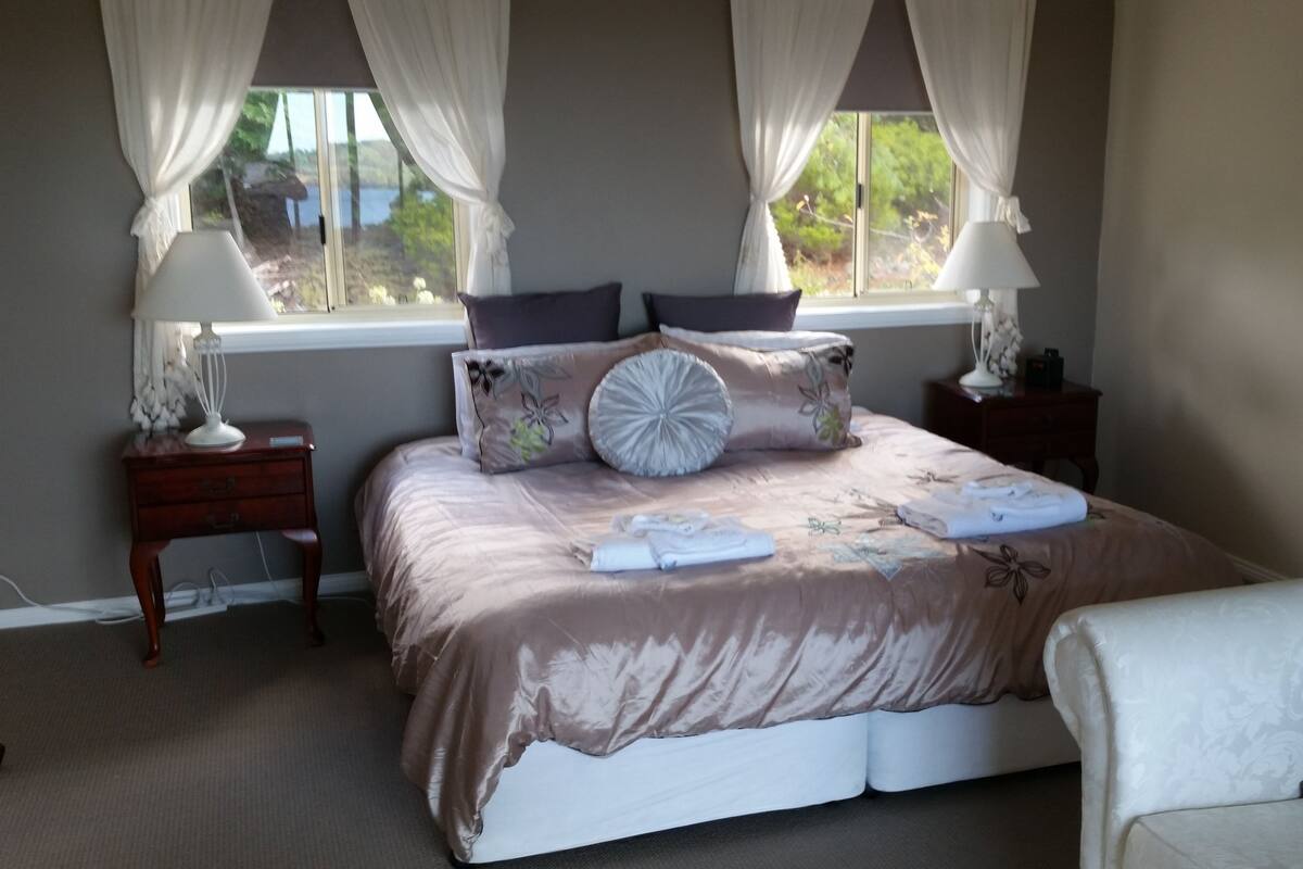 A bedroom with a large bed, two bedside tables with lamps, a window with curtains, and a view of greenery outside.