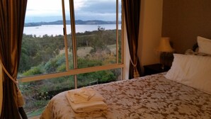 A bedroom with a large window offering a view of a lake and hills. There is a neatly made bed with a patterned bedspread and a folded towel on it. - Waterview Gardens B&B (Margate)