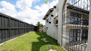 A courtyard with a wooden fence, a white building with a balcony, and a metal gate. - Motel Villa Del Rio (Whangarei)
