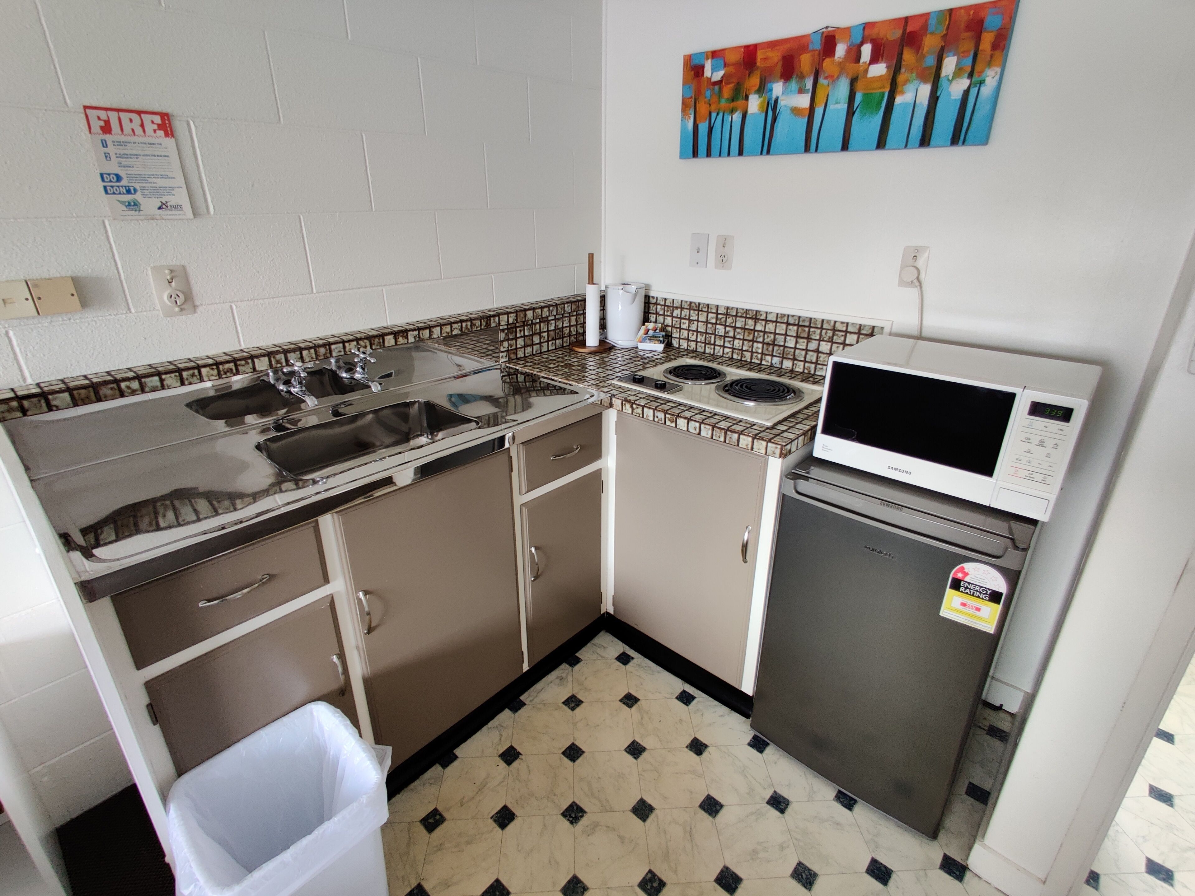 A compact kitchen with a microwave, stove, sink, and a trash bin.