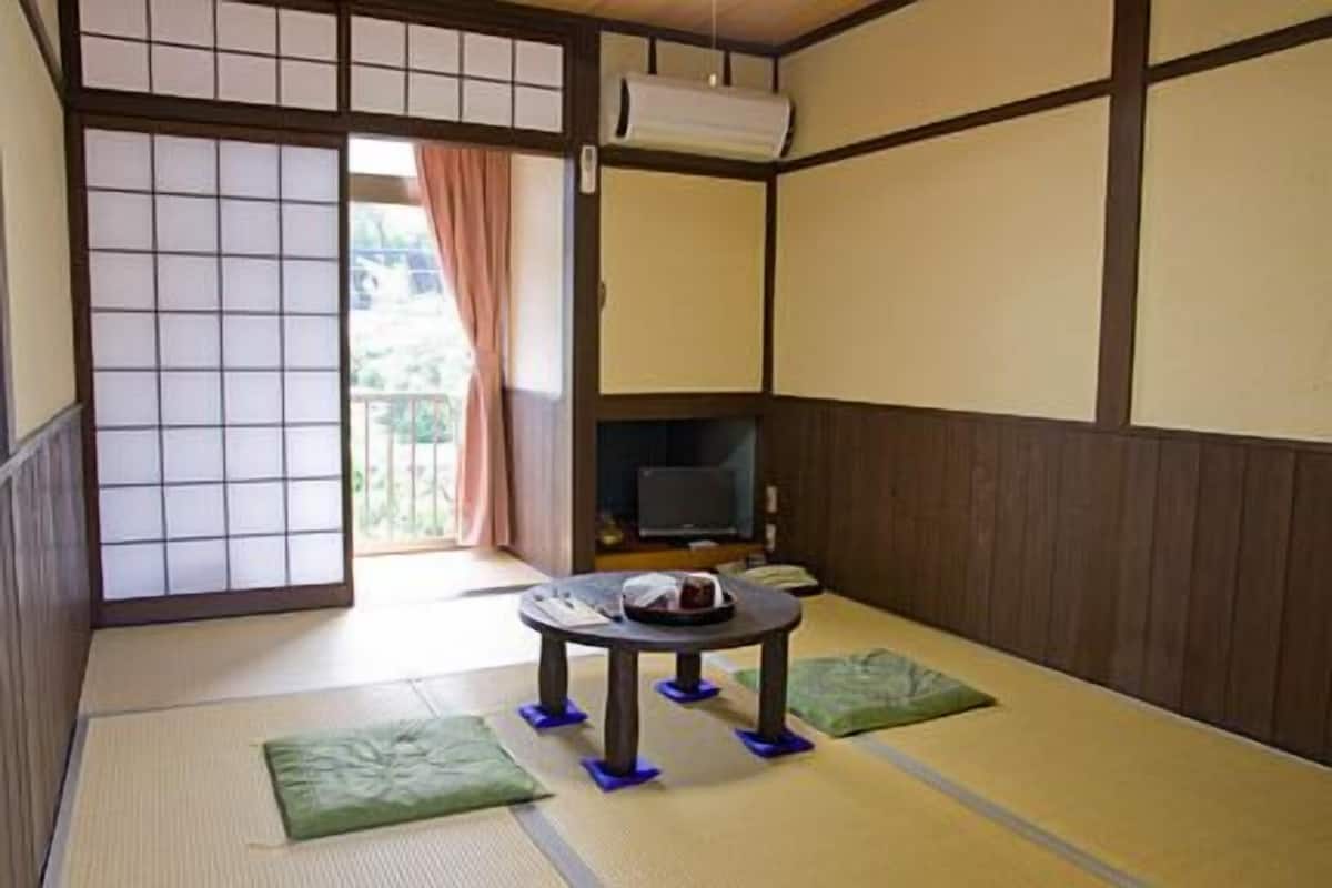 Une pièce de style japonais traditionnel, avec un sol recouvert de tatamis, une table basse en tatami, un téléviseur et une porte coulissante donnant sur un espace extérieur.