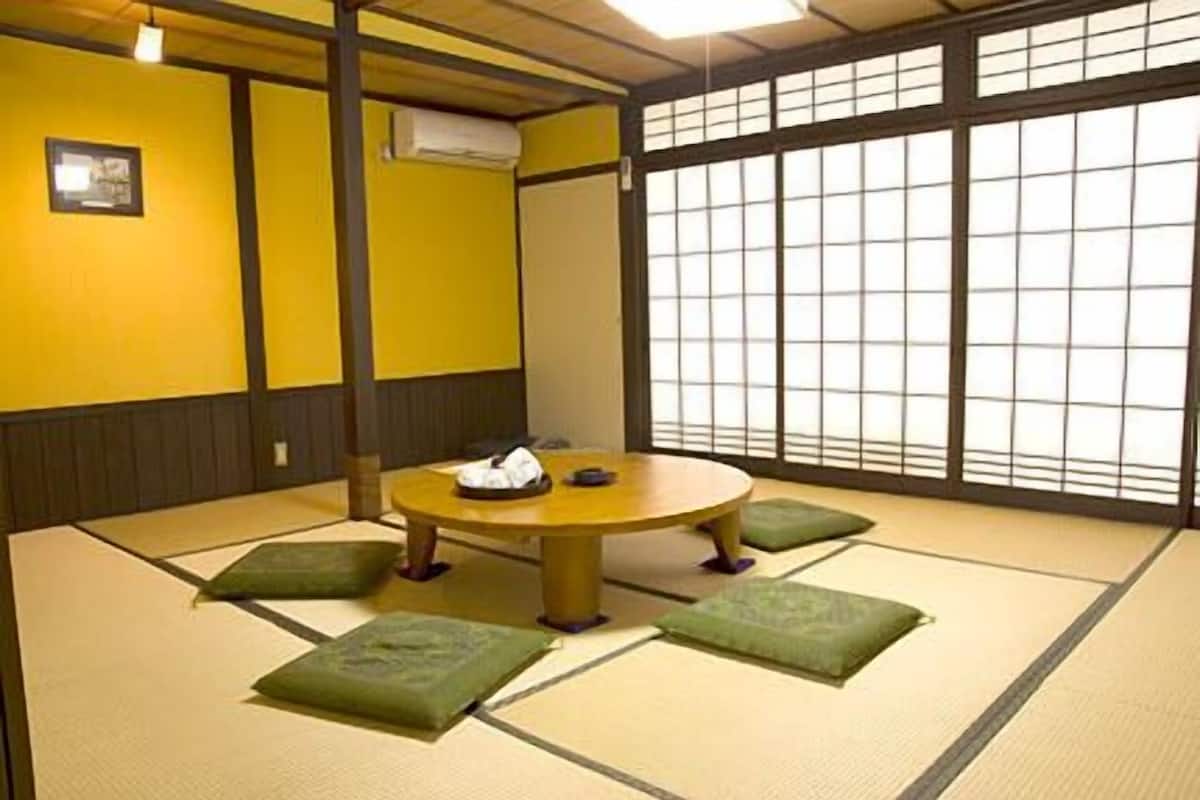 Une pièce avec un sol recouvert de tatamis, comprenant une table ronde, des coussins et un paravent de type shoji.
