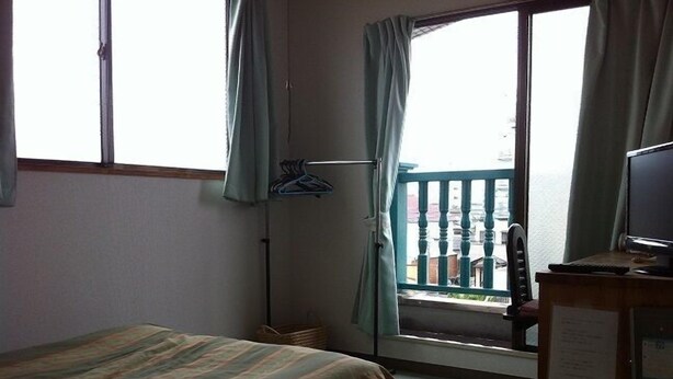 A hotel room with a bed, a desk with a computer, a window with curtains, and a view of buildings outside. - Business Hotel Mayumi (Omuta)