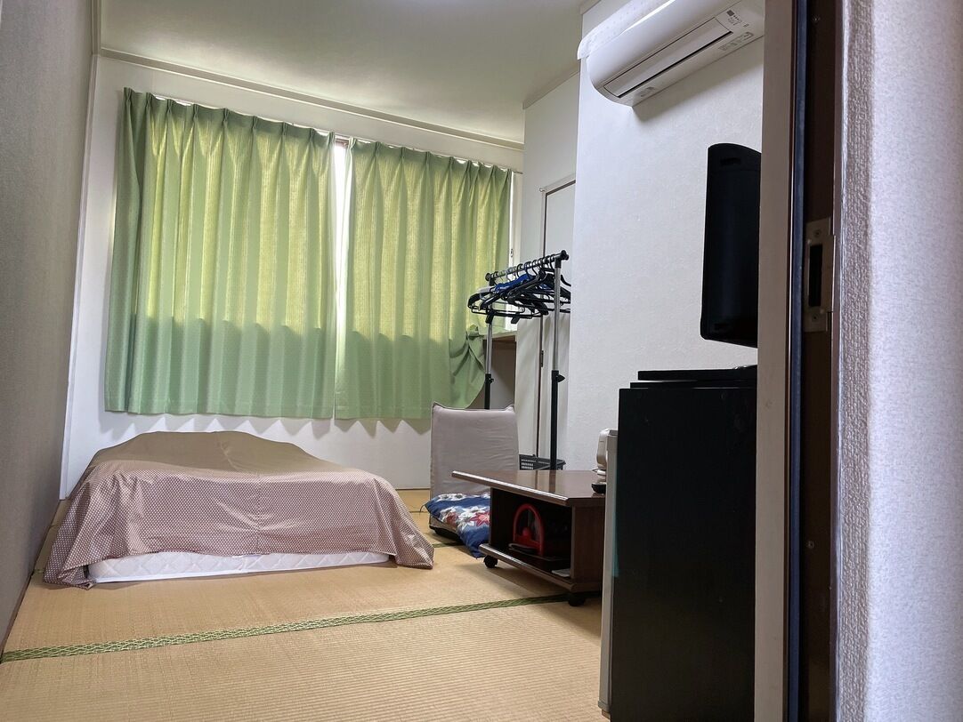 A small hotel room with a bed, a desk, a chair, a TV, and an air conditioning unit.