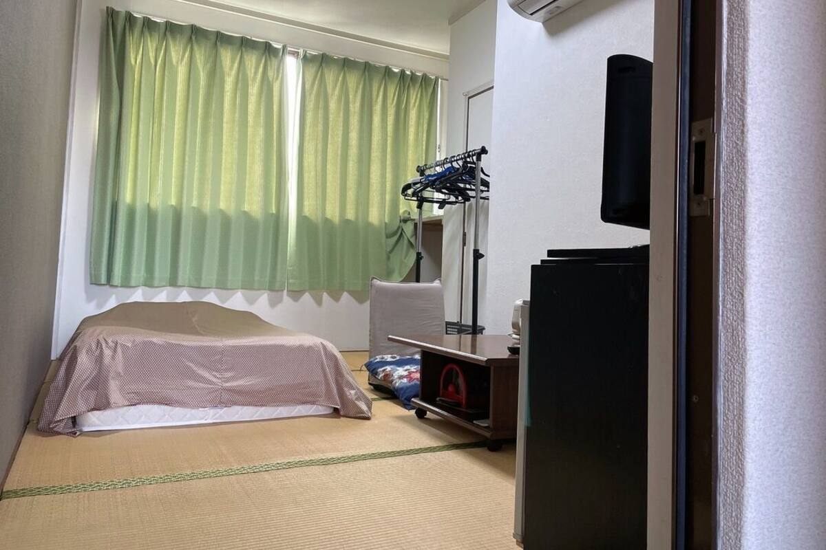 A small hotel room with a bed, a desk, a chair, a TV, and an air conditioning unit.