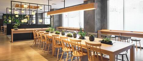 Un restaurante moderno con mesas y sillas de madera, plantas en macetas y un amplio ventanal con persianas.