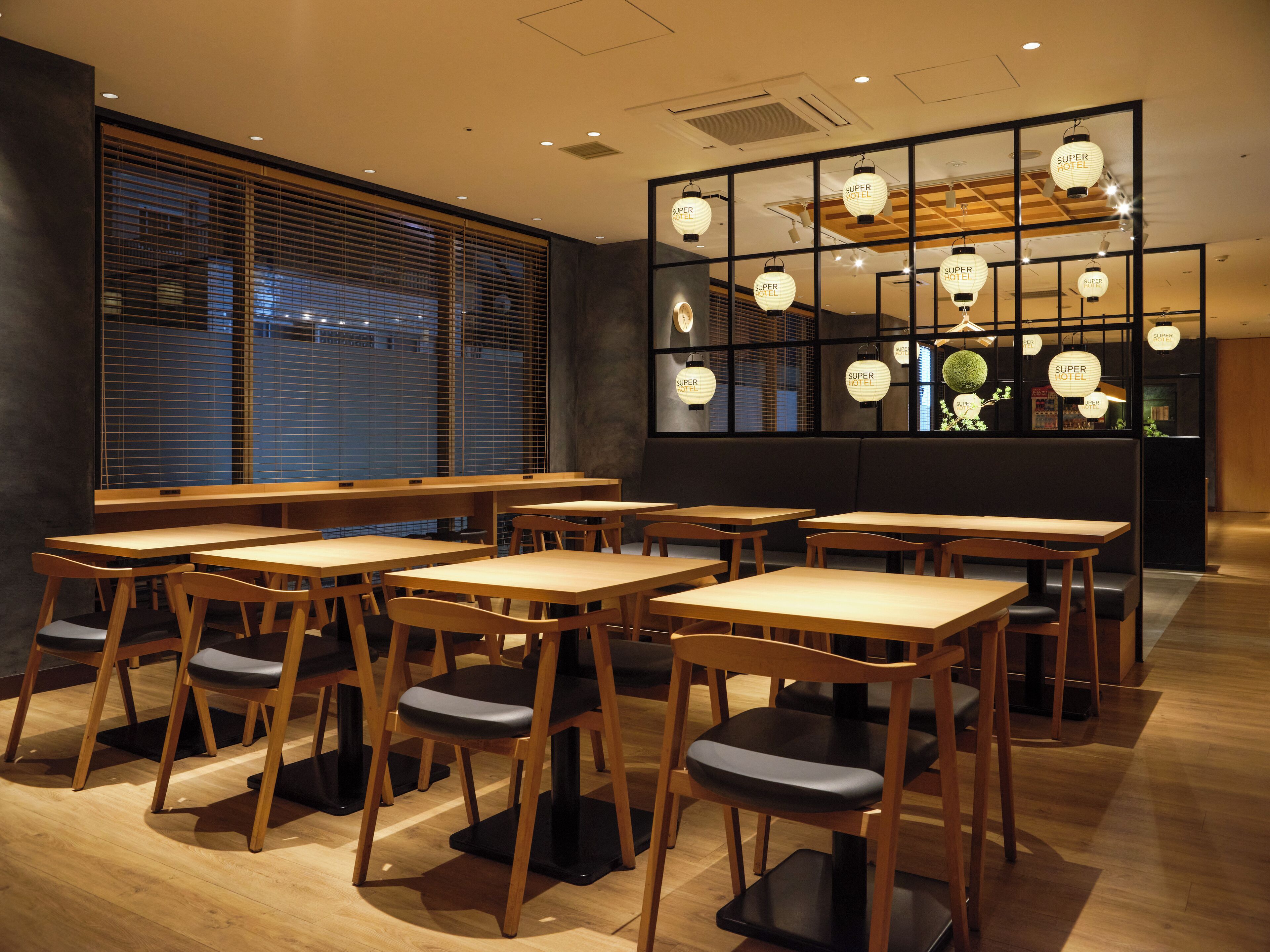 A restaurant with wooden tables and chairs, a large window with blinds, and hanging lanterns.