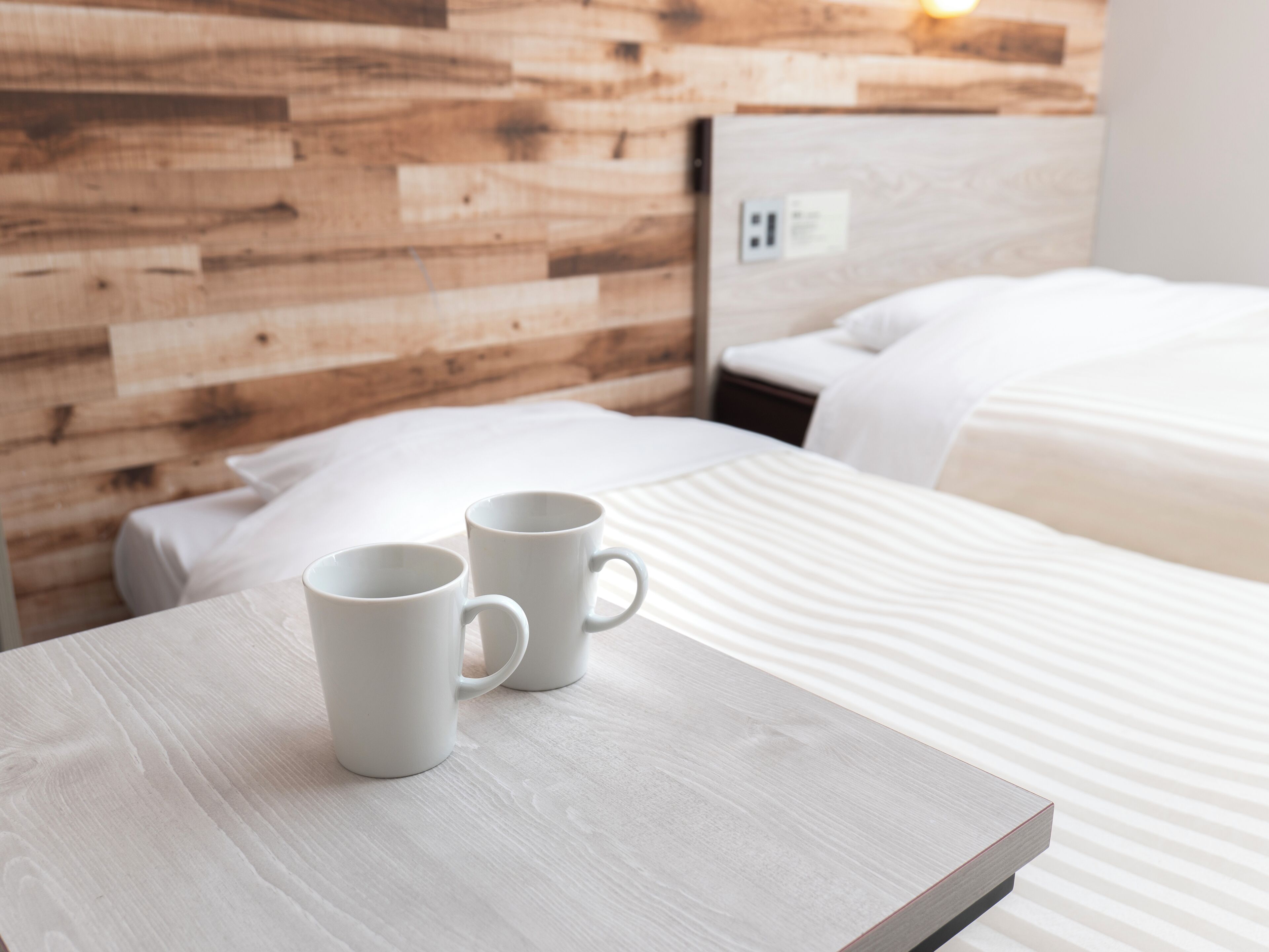 Ein ordentlich bezogenes Bett mit weißer Bettwäsche, ein hölzernes Kopfbrett und zwei weiße Tassen auf einem Holztisch.