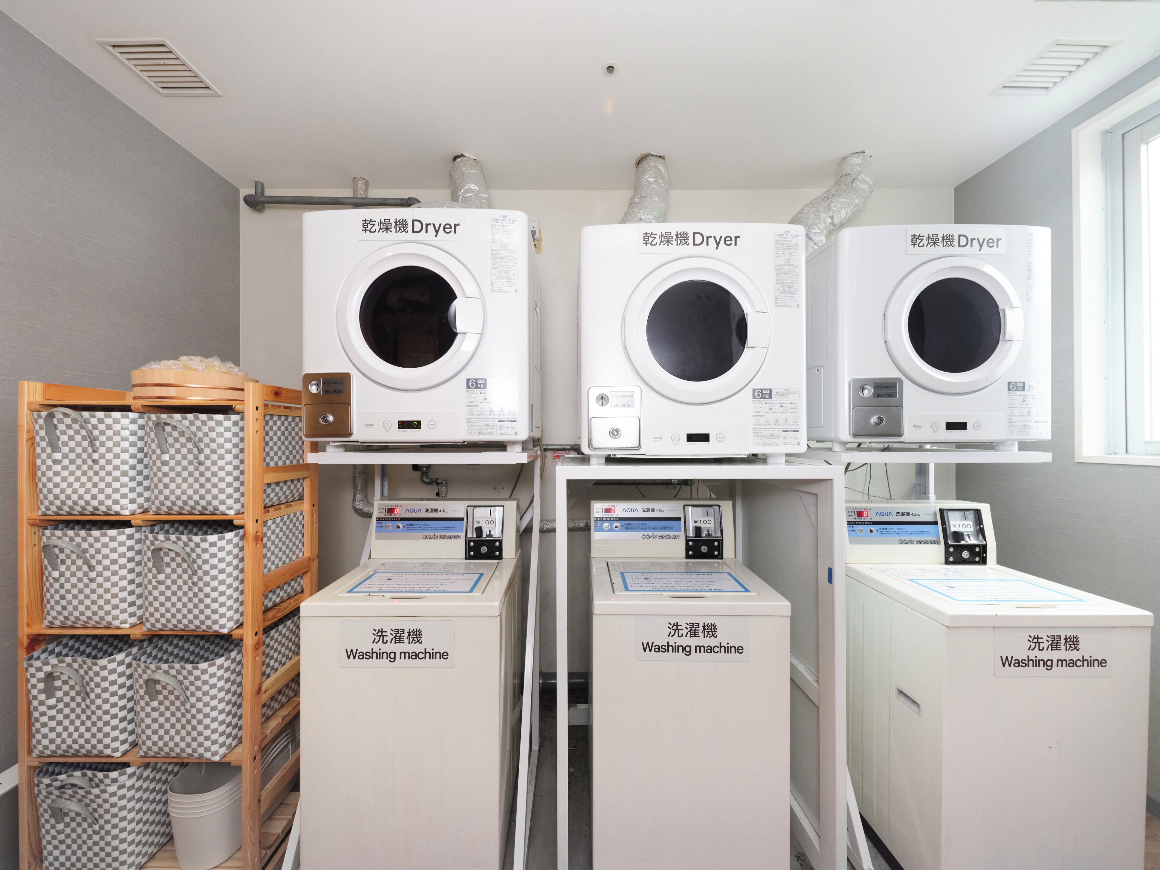 Three dryers and three washing machines in a laundry room with a wooden shelf holding checkered bags.