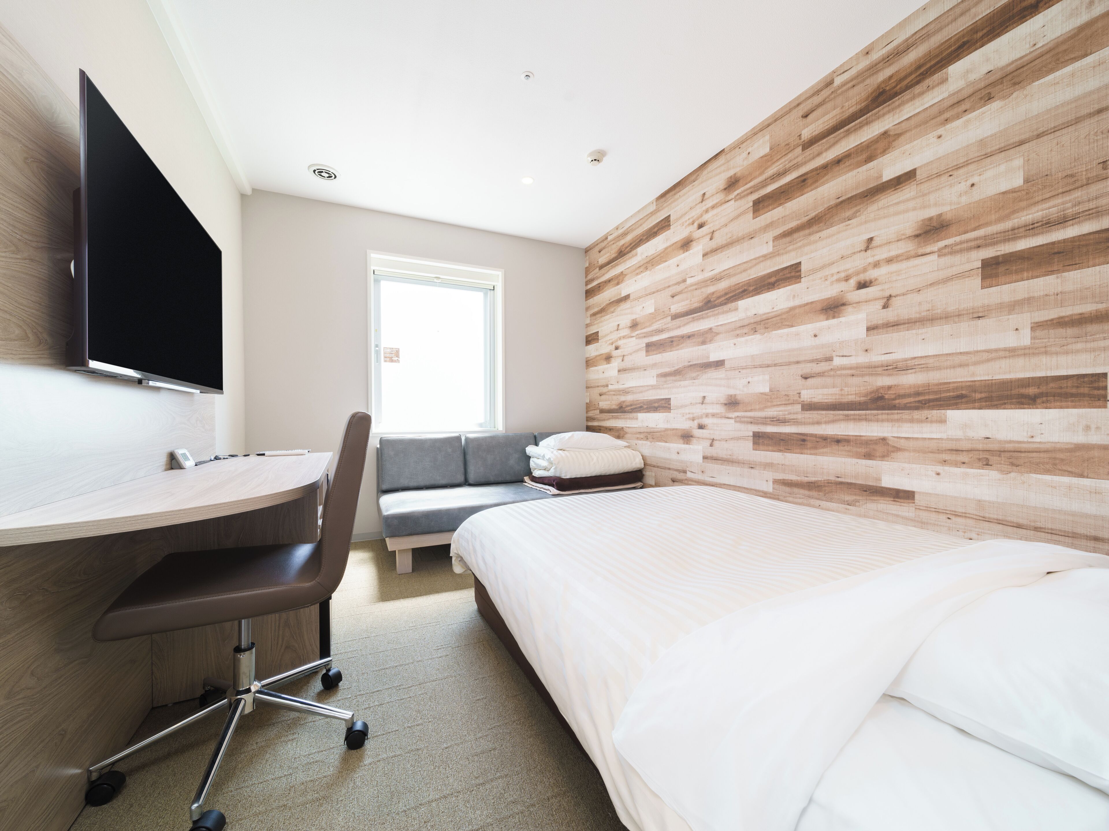 A modern hotel room with a wooden accent wall, a bed, a desk with a chair, a sofa, and a flat-screen TV mounted on the wall.